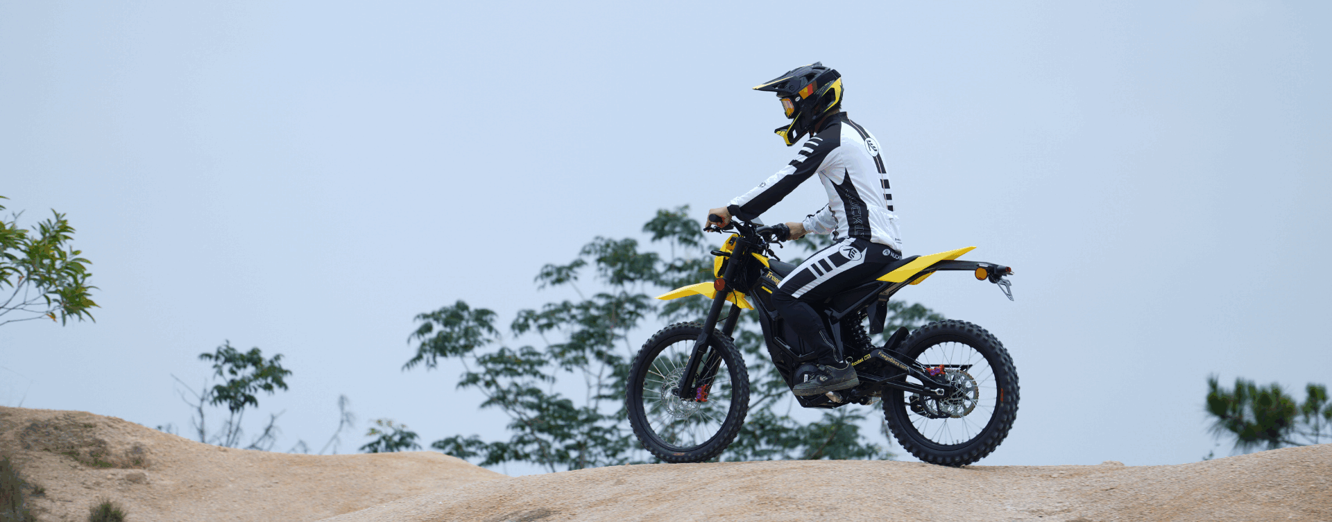Person riding a dirt bike on a sandy hill with trees in the background